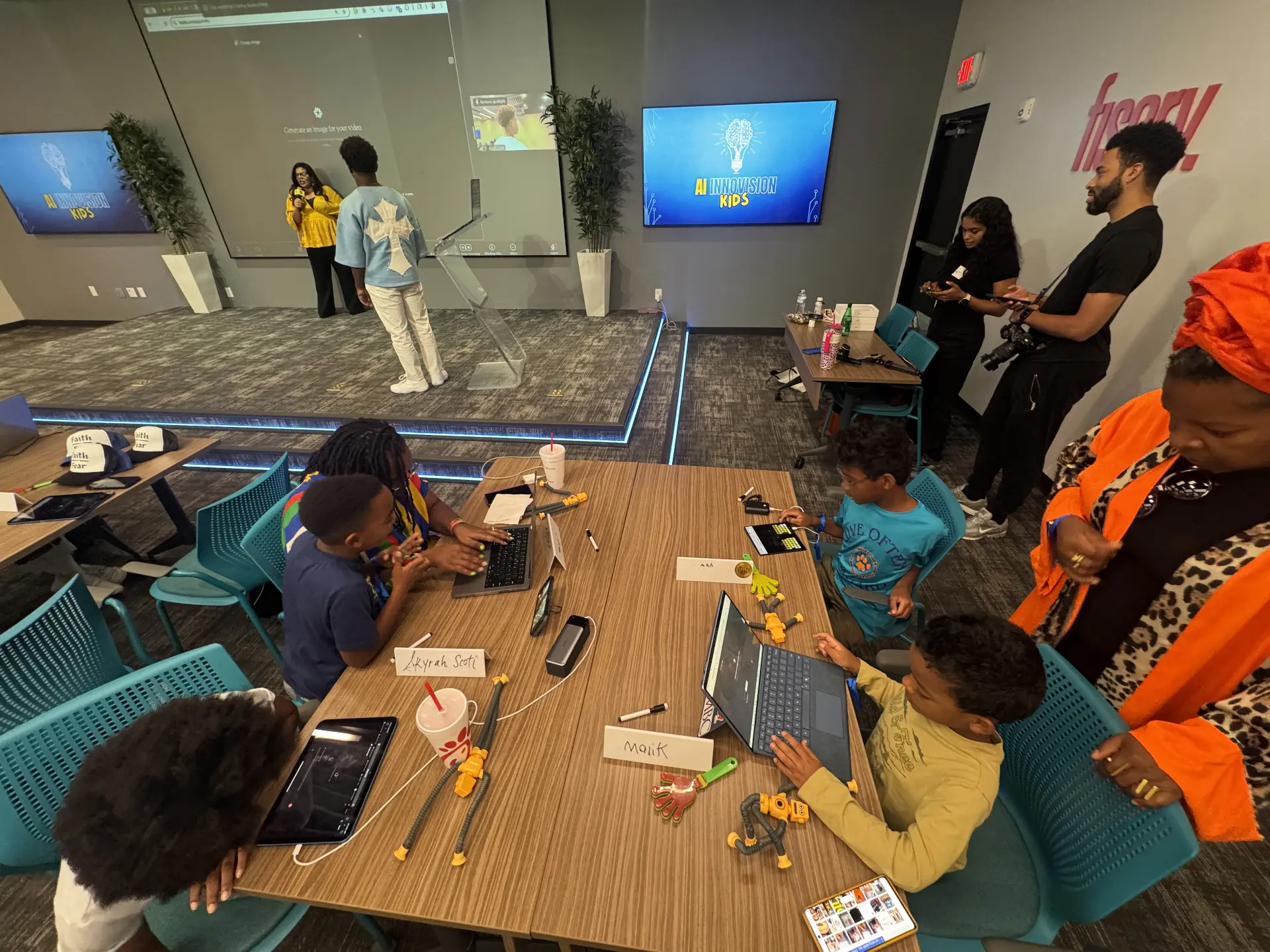 Children concentrating on laptops and tablets around a workshop table while adults assist nearby